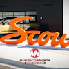 Scout Motors orange brand signage displayed outside showroom with tan SUV and Manufacturing Now logo