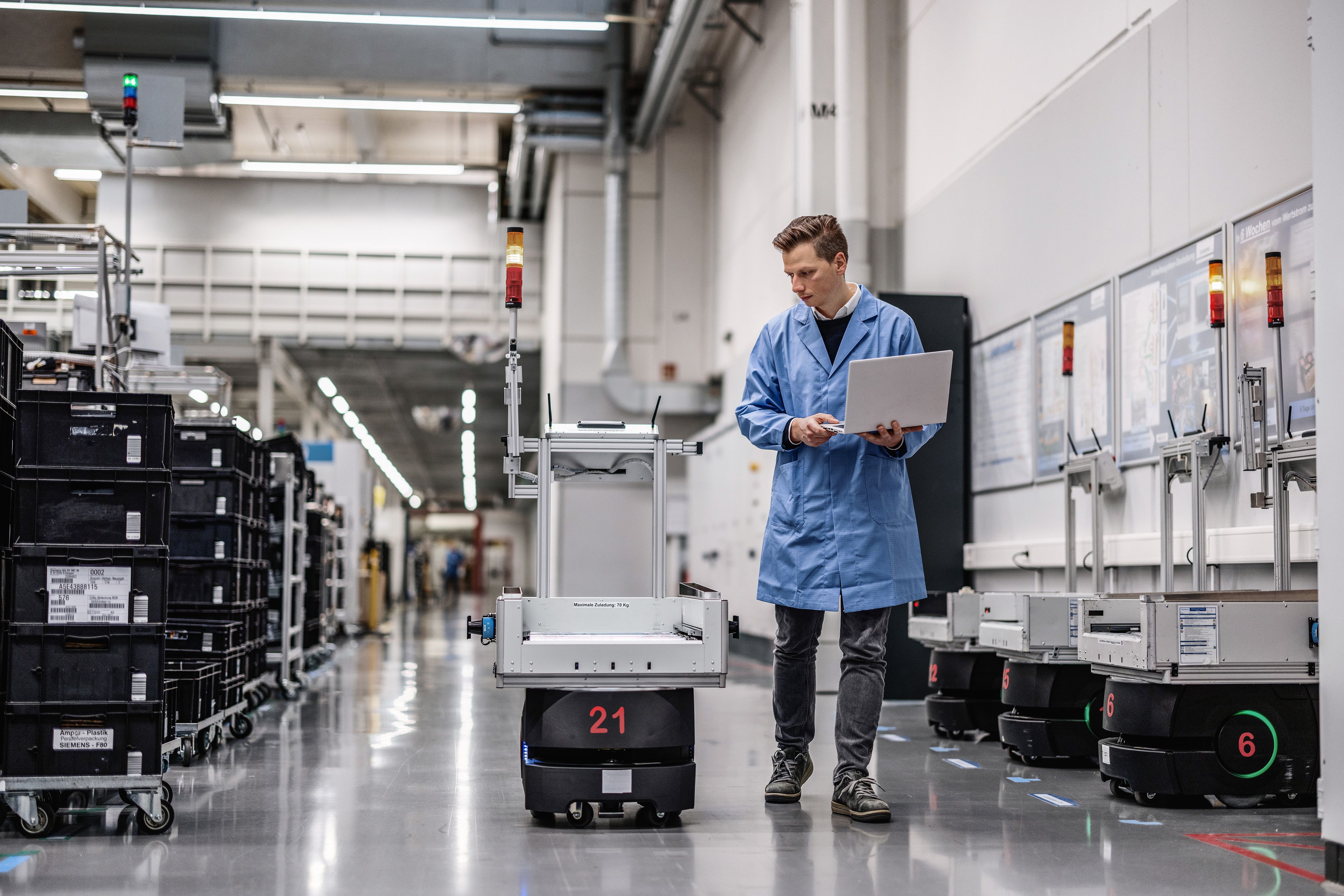 Technician in blue coat operating laptop beside autonomous mobile robot in industrial warehouse facility