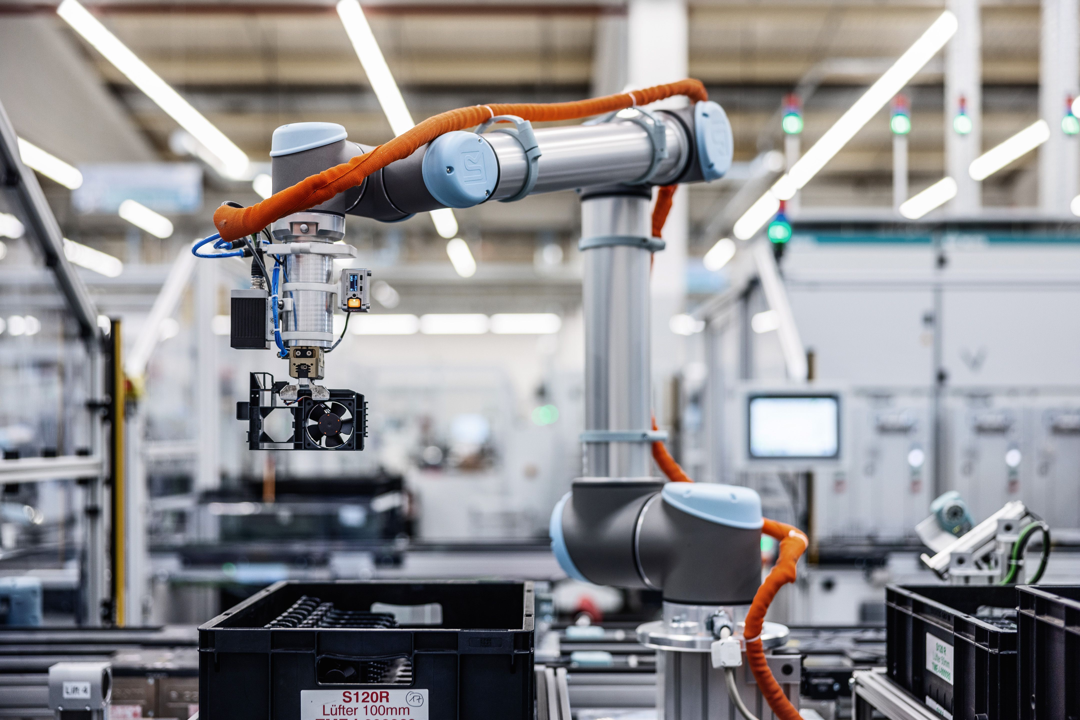 Two orange and silver robotic arms working together in a modern manufacturing facility with white structural elements and industrial equipment.