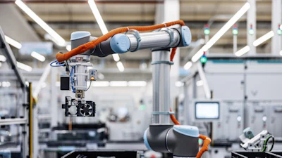 Two orange and silver robotic arms working together in a modern manufacturing facility with white structural elements and industrial equipment.