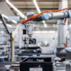 Two orange and silver robotic arms working together in a modern manufacturing facility with white structural elements and industrial equipment.
