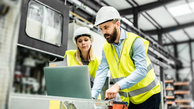 Two construction workers in hard hats and yellow safety vests review plans on a laptop inside an industrial warehouse facility.