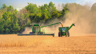 Green combine harvesters and tractors harvesting grain in a dusty field with trees in background