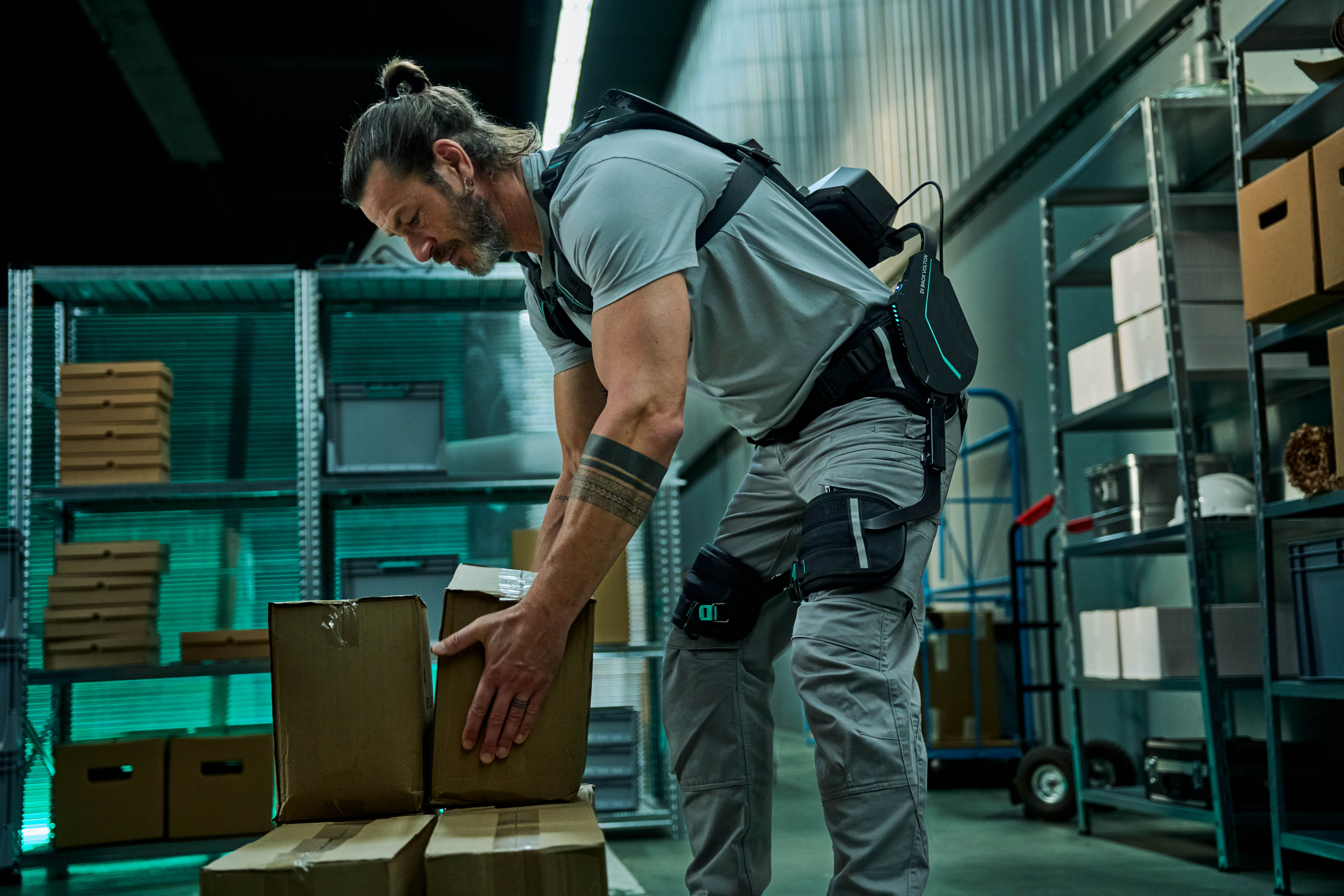 Worker in gray shirt and tool belt organizing cardboard boxes in a modern warehouse with teal shelving.