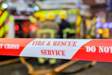 Red and white fire and rescue service barrier tape with 'Do Not Cross' warning at emergency scene with vehicles in background