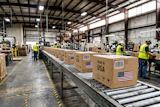 Workers in safety vests process cardboard boxes marked 'Made in USA' on a warehouse conveyor belt system.