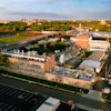 Aerial view of brick industrial complex with parking lot, green spaces, and surrounding cityscape