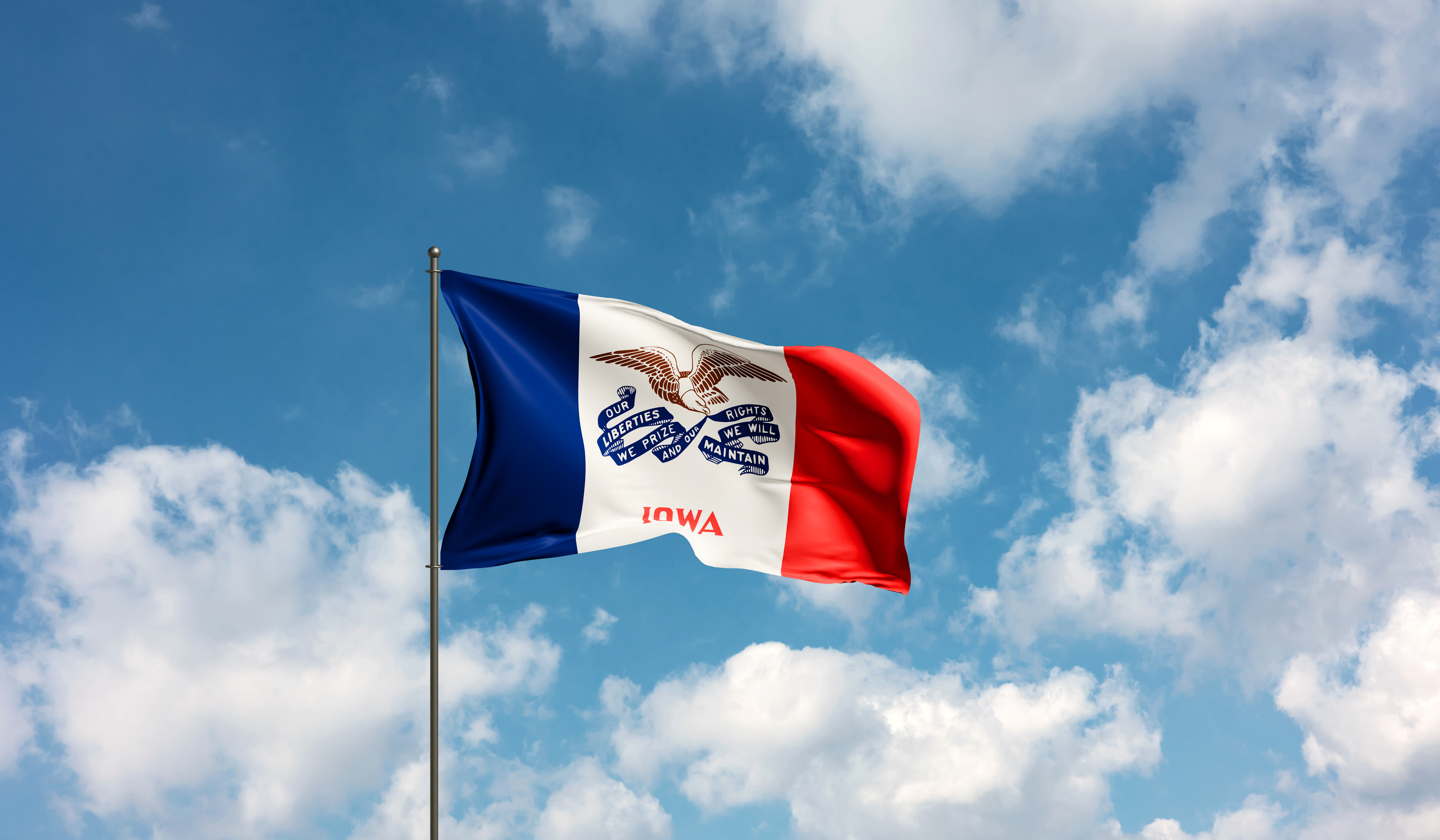 Iowa state flag waving on a flagpole against a blue sky with white clouds