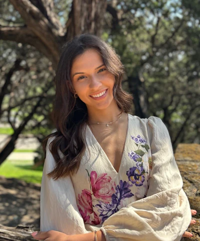 Woman smiling outdoors wearing floral white blouse under tree shade