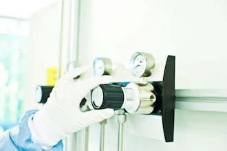 Gloved hand holding a brass gas regulator valve with pressure gauges and black control knob in laboratory setting