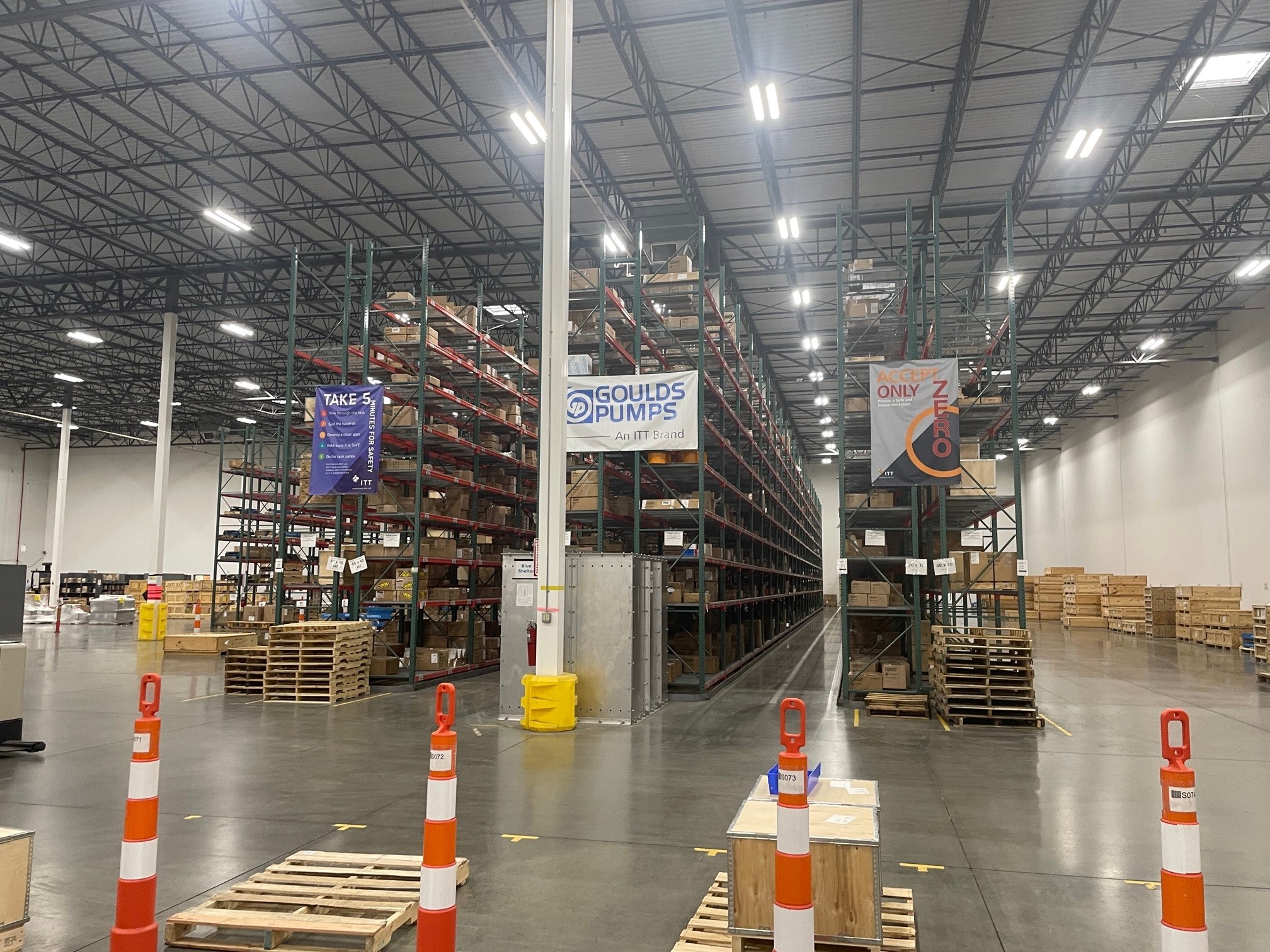 Industrial warehouse interior with tall storage racks, branded signage including Goulds Pumps and Rexnord logos, pallets, and orange safety markers on concrete floor.