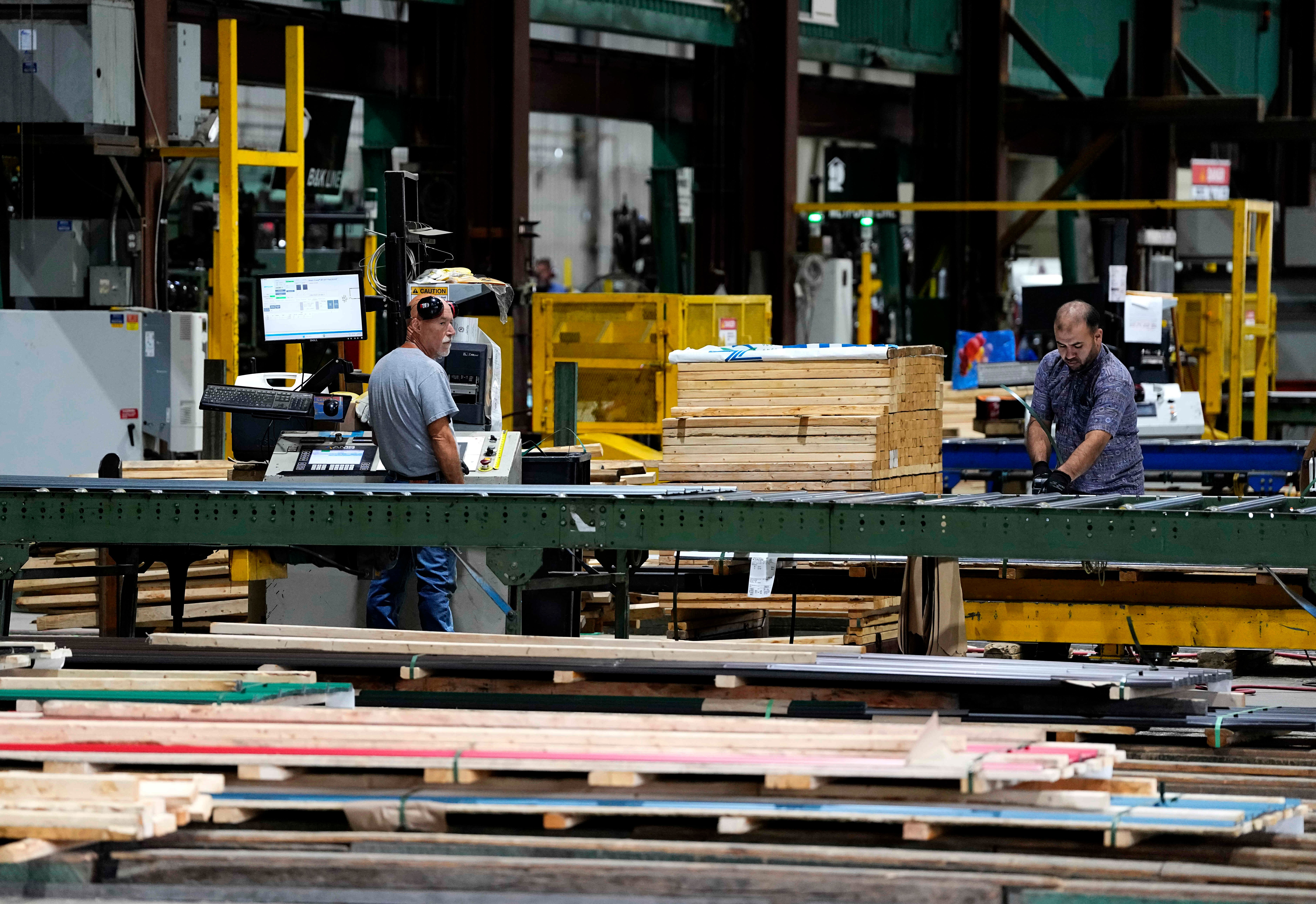 Workers processing lumber at an industrial woodworking facility with yellow machinery and green structural beams.