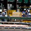 Workers processing lumber at an industrial woodworking facility with yellow machinery and green structural beams.