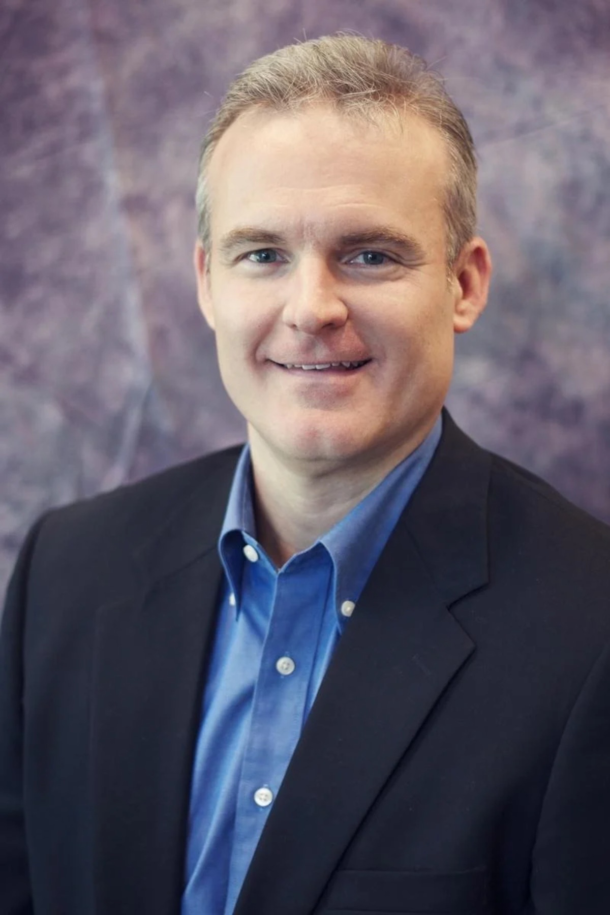 Professional headshot of a man in a dark blazer and blue button-up shirt smiling at the camera
