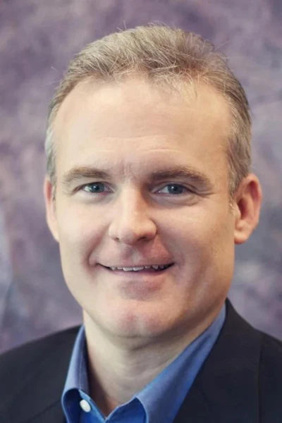 Professional headshot of a man in a dark blazer and blue button-up shirt smiling at the camera