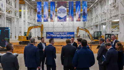 Corporate event in industrial warehouse with blue banner reading 'Future Home of 24 Hadrian Machines' and yellow excavators