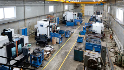 Industrial manufacturing facility with CNC machines, blue workbenches, and overhead yellow crane system in warehouse space