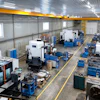 Industrial manufacturing facility with CNC machines, blue workbenches, and overhead yellow crane system in warehouse space