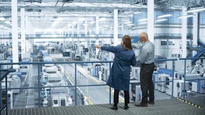 Two professionals observe a large industrial manufacturing facility from an elevated platform with expansive windows.