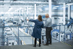 Two professionals observe a large industrial manufacturing facility from an elevated platform with expansive windows.