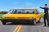 Man in black clothing posing beside a bright yellow vintage minivan on a rural highway with green fields and blue sky