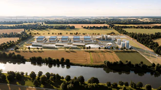Aerial view of industrial facility with blue-roofed buildings, storage tanks, and surrounding agricultural fields near a river