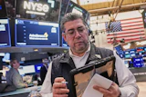 Trader at NYSE trading floor wearing headset and holding tablet with monitors displaying market data and American flag in background
