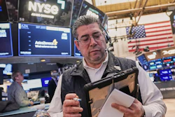 Trader at NYSE trading floor wearing headset and holding tablet with monitors displaying market data and American flag in background
