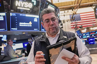 Trader at NYSE trading floor wearing headset and holding tablet with monitors displaying market data and American flag in background