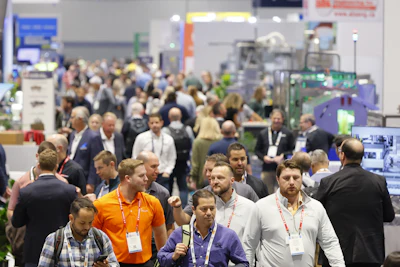 Crowded trade show floor with attendees wearing lanyards and ID badges networking among exhibition booths