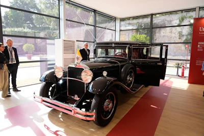 Vintage black automobile from the 1920s-30s displayed in a modern museum gallery with large windows and visitors observing the classic car.