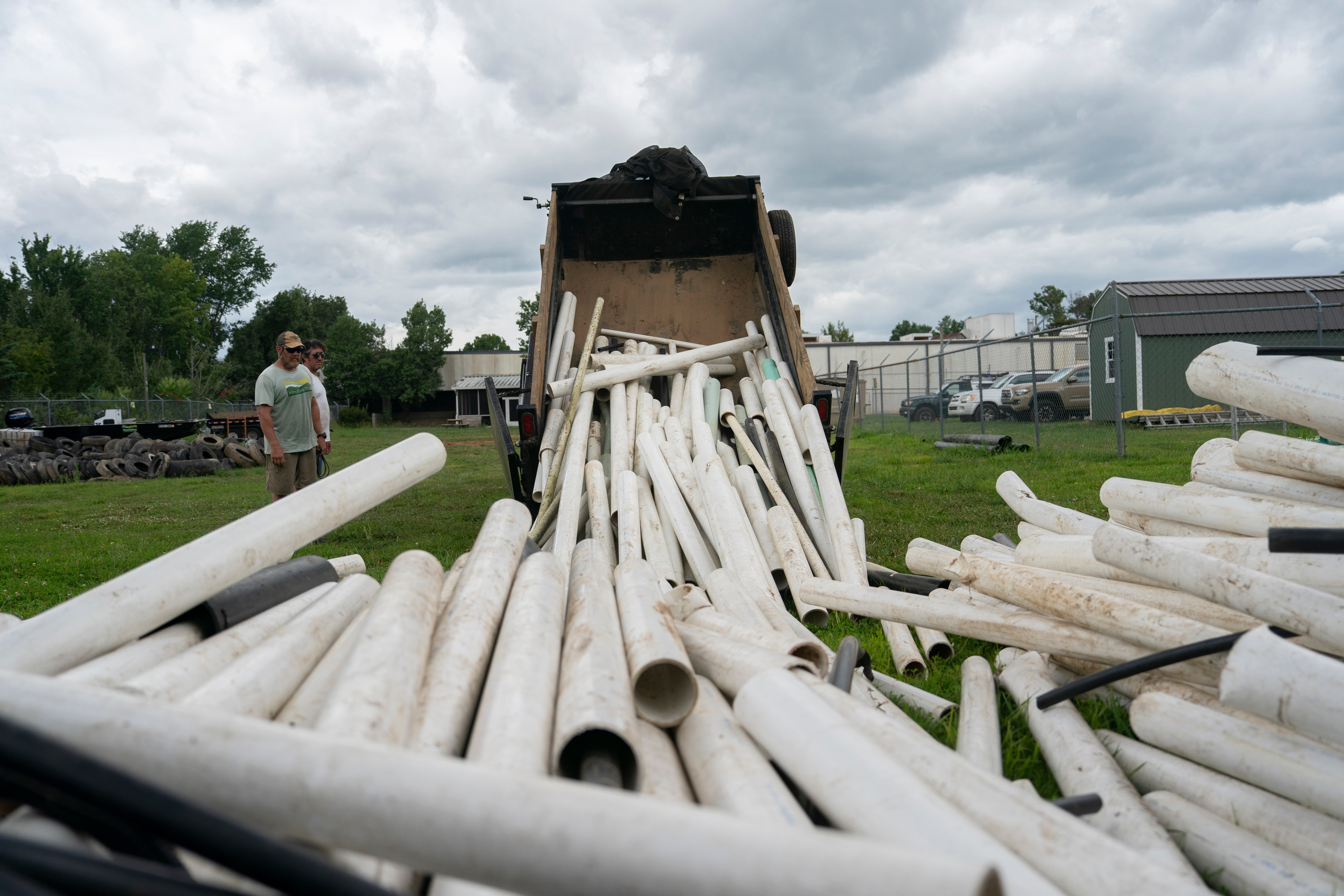 White PVC pipes being unloaded from a dump truck at an industrial yard with green buildings in background