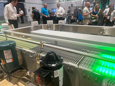 Industrial conveyor belt system with green LED lighting at manufacturing facility or trade show event with visitors observing equipment.