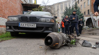 Damaged BMW sedan with detached engine component displayed on ground during investigation with officials and onlookers in urban courtyard