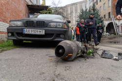 Damaged BMW sedan with detached engine component displayed on ground during investigation with officials and onlookers in urban courtyard