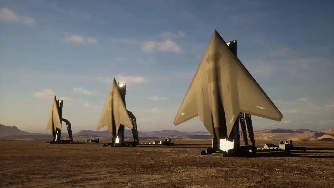 Three large metallic sail-shaped sculptures stand in a desert landscape under a blue sky with clouds