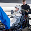 Astronaut in white SpaceX suit waving while seated in spacecraft with crew members assisting
