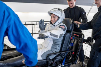 Astronaut in white SpaceX suit waving while seated in spacecraft with crew members assisting