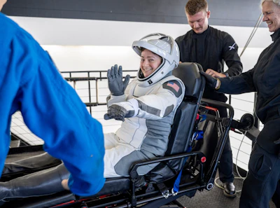 Astronaut in white SpaceX suit waving while seated in spacecraft with crew members assisting