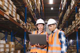 Two warehouse workers in safety gear reviewing inventory data on laptop in storage facility