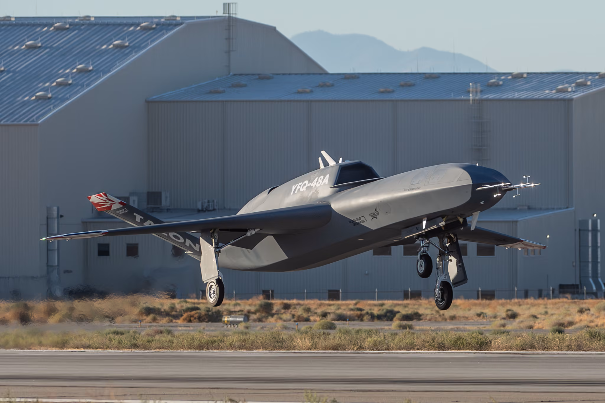 Silver and black experimental aircraft landing near industrial hangar with mountains in background