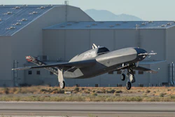 Silver and black experimental aircraft landing near industrial hangar with mountains in background