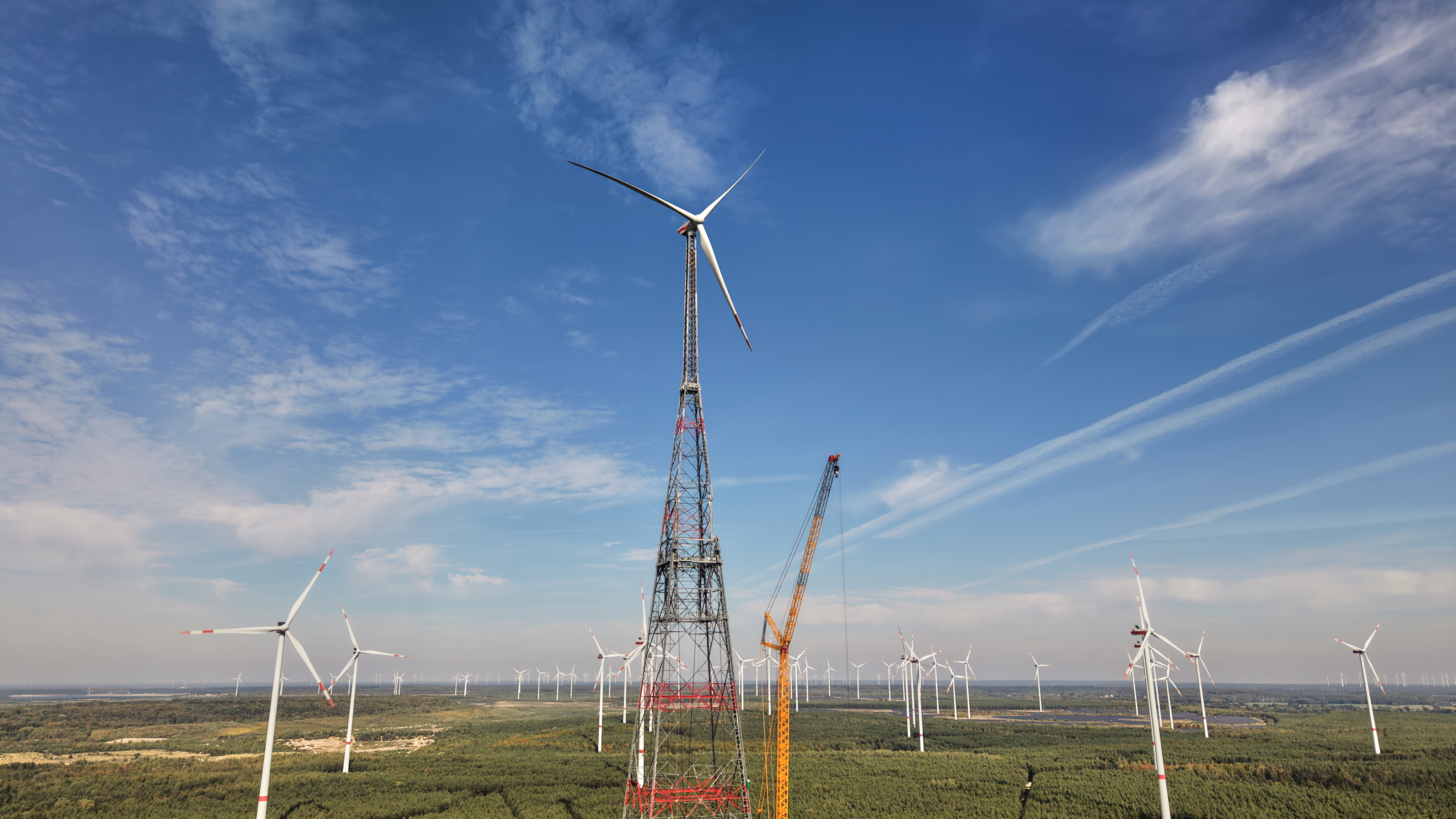 Wind farm with red and white lattice tower and multiple turbines under blue sky